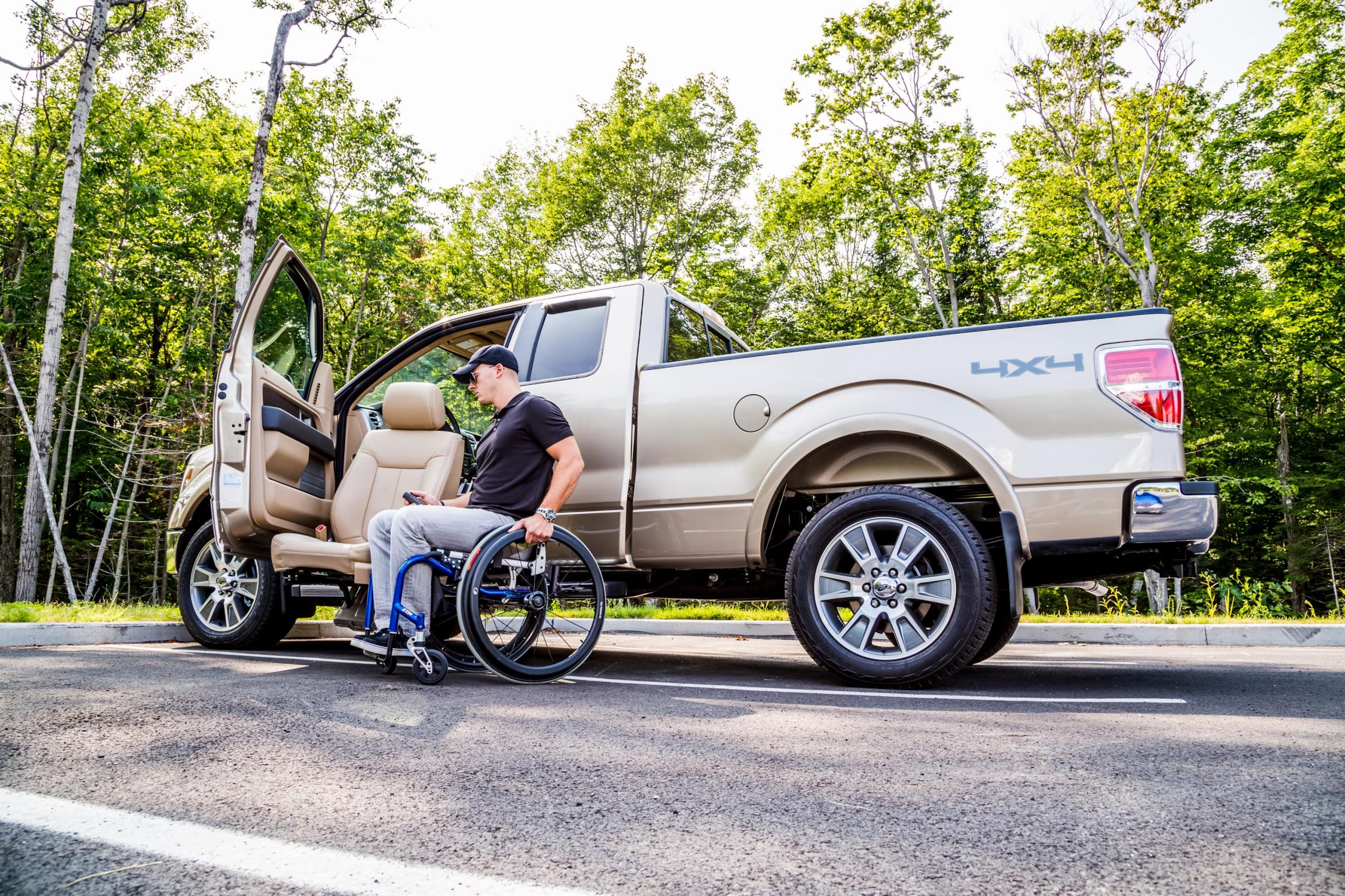 A man sits in a rigid frame wheel chair outside a tan Ford pickup truck. The OEM seat is outside the vehicle on the LINK.